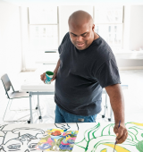 Jimmy stands in a relatively empty room with a large canvas in front of him. He holds a cup of paint in one hand and a paintbrush in the other