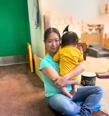 Mei-Feng sits cross legged on the floor of daycare while holding a baby
