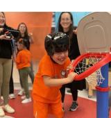 Student hangs on a small basketball hoop, he has a big grin on his face and there are many people around him smiling and cheering him on