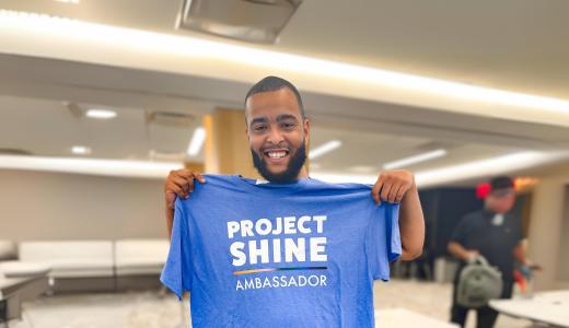 Breon smiles and poses for photo holding up a project SHINE t-shirt