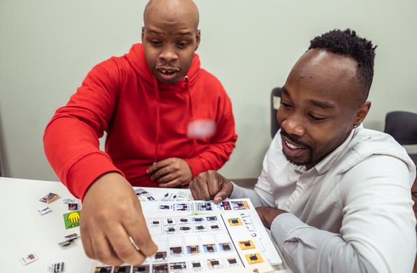 Two men sit at at table, one on left wears red, one on right wears white, they appear to be organizing small images