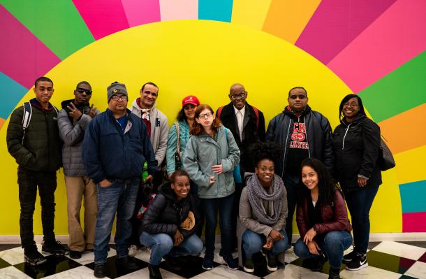 Large group of people supported by YAI and staff standing and kneel in front of a very colorful sun with multicolor rays for a group photo