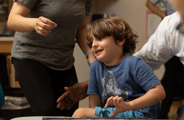 A child around age 7 is sitting at a table, you can see two adults by the child, they appear to be helping them.