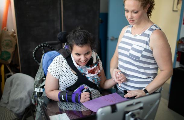 iHOPE student is in a wheelchair and looking at a tablet and is drawing on paper with support from therapist holding their hand