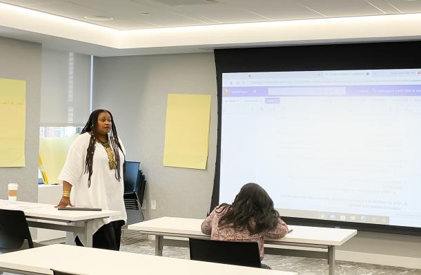 Consuelo Senior stands to the left by a table and powerpoint on the right, she is providing a training