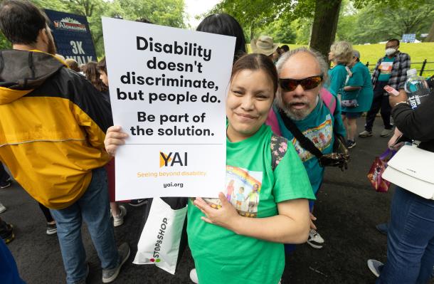 two people in a crowd at central park challenge, one holds a sign "Disability doesn't discriminate, but people do. Be part of the solution"
