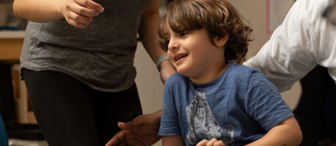 A child around age 7 is sitting at a table, you can see two adults by the child, they appear to be helping them.