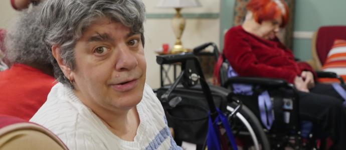 Person with shorter grey hair and sweater is sat down looking at camera to their right. In the background you can see another person in a wheelchair and possibly 2 other people behind the person in the foreground