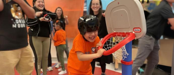 Student hangs on a small basketball hoop, he has a big grin on his face and there are many people around him smiling and cheering him on