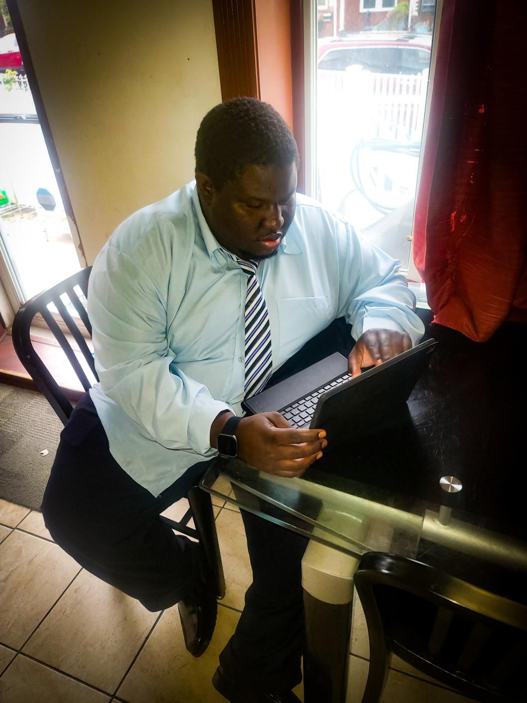 Person sits at a table looking at a laptop on the table. They are wearing a light blue button down shirt and tie