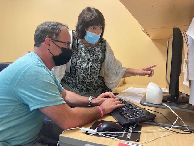 Two people at a computer, one is sat typing at the keyboard, the other is stood nearby pointing at the monitor.