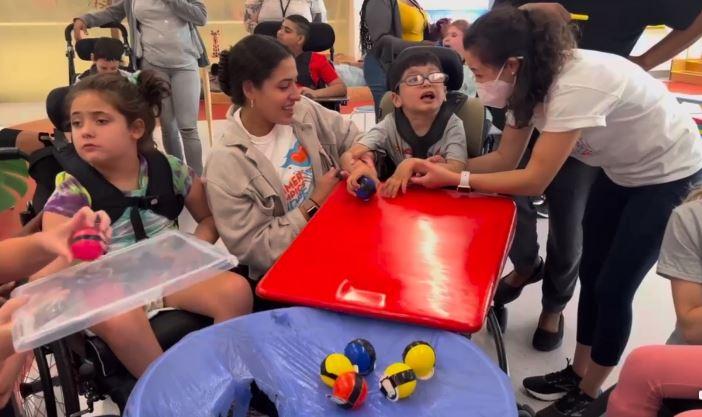 Two adults support an elementary school aged child in a wheelchair with holding and dropping a toy penguin