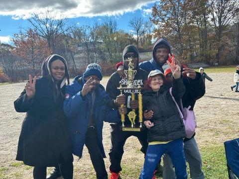 Group of 5 people stand in a field around a large trophy