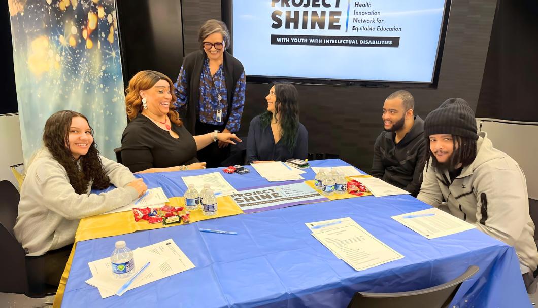 5 people sit at a table, one person is stood by them. You see the Project Shine logo on the screen in the background