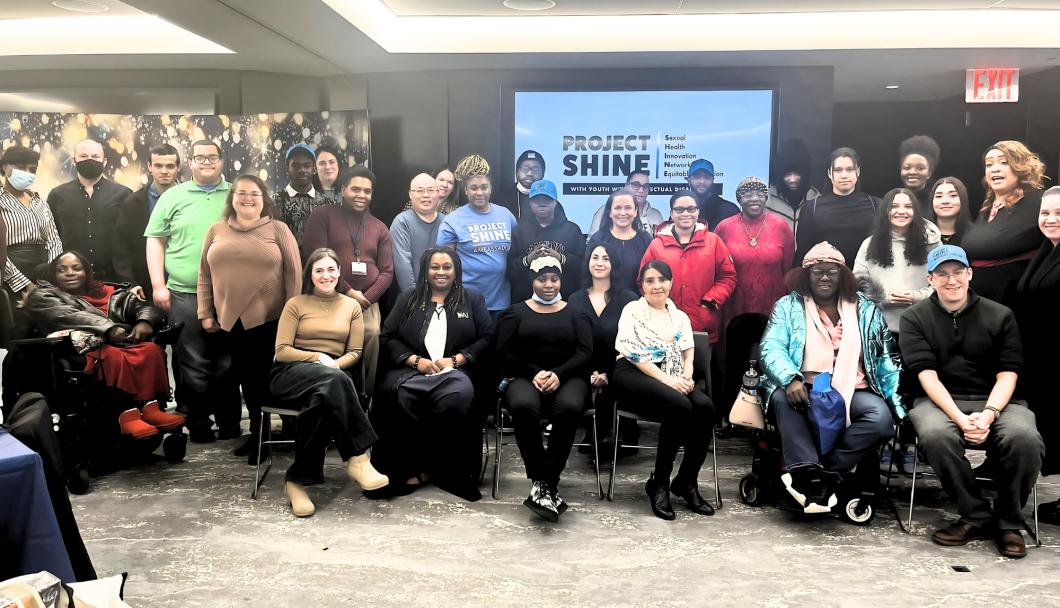 Large group of more than 20 people sit and stand in a posed group shot. You see a screen with "project SHINE" in the background