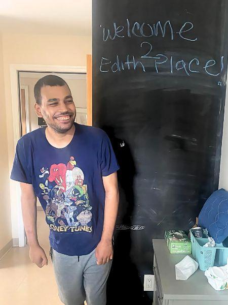 Roberto stands and is smiling next to a chalkboard with "Welcome to Edith Place" written on it