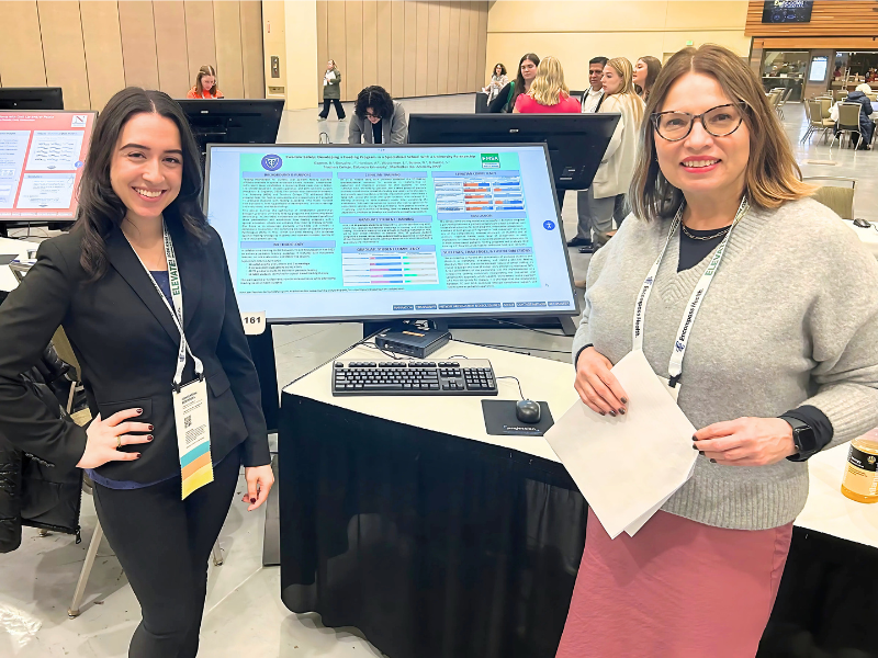 Two women stand in front of a large monitor at a conference