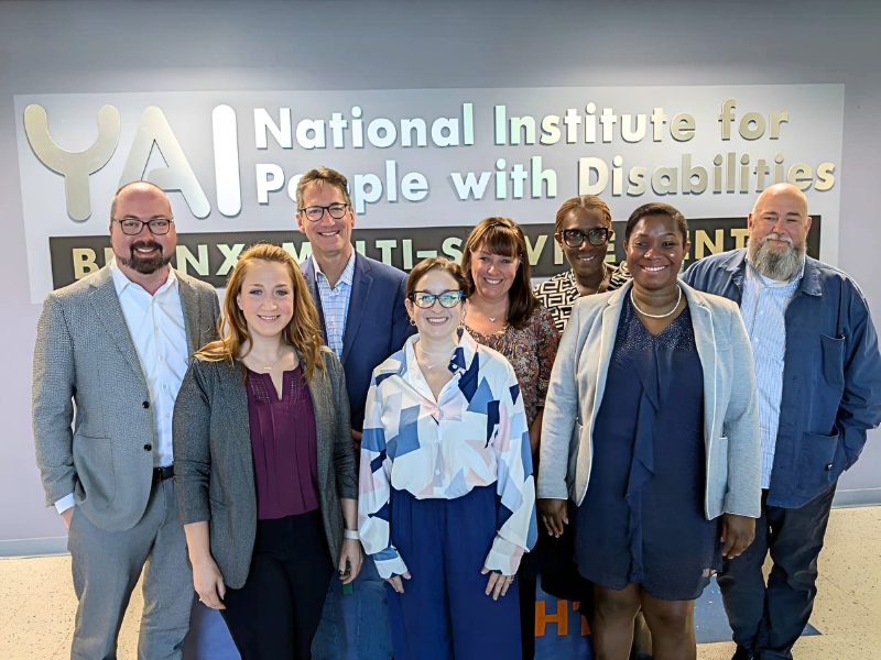 Group of 8 people pose for photo in front of the YAI sign