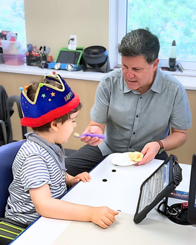 Young boy sits at a table, a man to his left has a fork with cupcake on it going into his mouth